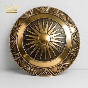 May include: A circular gold-toned shield featuring a prominent central sunburst design with radiating rays. The outer border is decorated with geometric triangular patterns. The surface has a metallic finish with dark recessed detailing.
