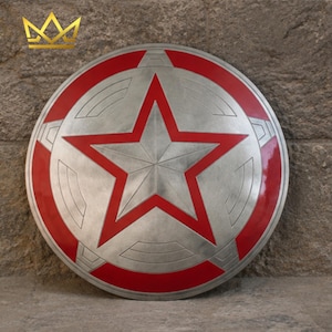 May include: A circular shield featuring a metallic silver finish with a prominent central five-pointed star outlined in red. The design includes concentric circular grooves and red accents along the outer rim, creating a geometric pattern.