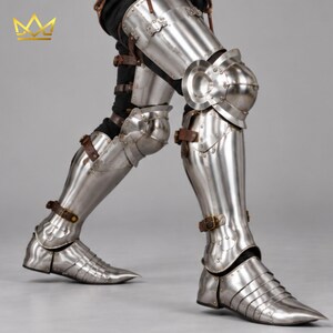 May include: Polished steel leg armor featuring articulated thigh plates, knee cops, greaves, and pointed sabatons. The metal components are secured with brown leather straps and buckles, showing a metallic silver finish with visible rivets throughout.