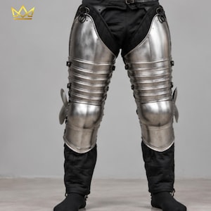 May include: A pair of articulated steel leg armour pieces featuring polished metal plates covering the thighs and knees, secured with side straps and rivets. The armour is worn over black quilted fabric trousers that extend down to the lower legs.