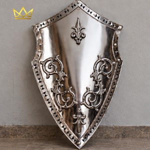 May include: A polished silver-coloured metal shield with a pointed heater shape. The surface features a central fleur-de-lis emblem, symmetrical scrollwork patterns on both sides, and a decorative border with small triangular cutouts and rivets.