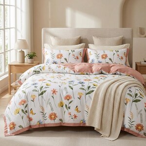 May include: A white duvet cover and matching pillowcases featuring a colourful botanical print of orange, yellow, and blue wildflowers with small yellow butterflies. The bedding set includes a solid dusty pink border and a beige knit throw blanket.