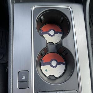 May include: Two circular car cup holder inserts featuring a red, white, and black graphic design resembling a Poké Ball. The coasters have a distressed, textured appearance and are positioned inside the dual cup holders of a vehicle console.