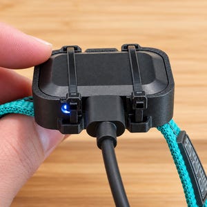 May include: A black rectangular electronic device attached to a bright teal woven strap with two black zip ties. A black charging cable is plugged into the center of the device, and a small blue LED light is illuminated on the side.