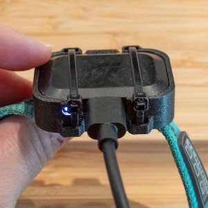 May include: A black rectangular electronic device attached to a teal fabric collar with black plastic zip ties. A black charging cable is plugged into the center of the device, and a small blue LED light is visible on the side of the housing.