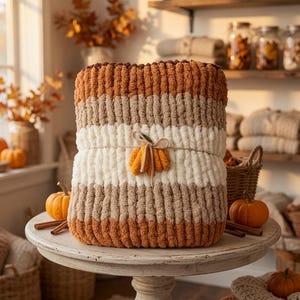 May include: A chunky knit throw blanket featuring horizontal stripes in shades of burnt orange, beige, and cream. The blanket is folded and secured with a piece of twine tied in a bow, which holds a small decorative orange yarn pumpkin.