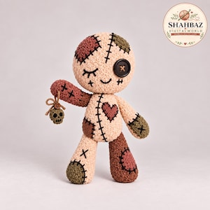 May include: A beige crochet voodoo doll featuring a black button eye, a stitched eye, and a heart patch. The doll has patches in olive green and maroon, black cross-stitch details, and holds a small crocheted skull charm by a brown string.