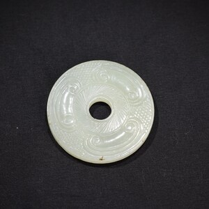 May include: A circular, off-white jade disc featuring a central hole and intricate, carved relief patterns. The surface displays swirling, cloud-like motifs and textured cross-hatching details arranged symmetrically around the center opening.