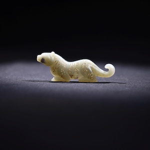 May include: A small, pale, off-white carved figurine of a feline animal in a crouching position. The surface of the figurine is decorated with intricate, dark, etched calligraphic characters along its body, head, and curved tail.