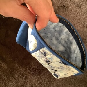 May include: A quilted fabric pouch featuring a blue and white floral pattern on the exterior and a light blue interior lining with a small white floral print. The pouch is secured with a dark metal zipper along the top edge and has a blue fabric trim.