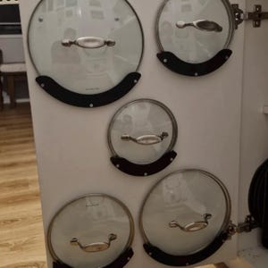 May include: Five circular glass pot lids with silver-toned metal handles are mounted on a white cabinet door. Each lid is held in place by a black, semi-circular plastic bracket secured with screws. The lids vary in size and feature the Scanpan logo.