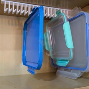 May include: A white plastic storage rack with vertical slots holds several square food container lids. The lids are various sizes and colors, including bright blue, light teal, and translucent white, each featuring locking side tabs.