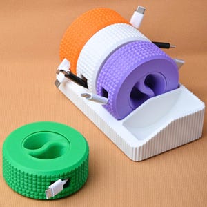 May include: A set of circular cable organizers with a textured grid pattern. Three organizers in orange, white, and purple are stacked in a white rectangular base, while a green organizer sits separately. Each unit features a central cutout.