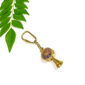 May include: A brass keychain featuring a miniature ship engine order telegraph with a circular face, a central anchor symbol, and a conical base. The telegraph dial displays text including FULL, HALF, SLOW, and DEAD SLOW in a dark red background.