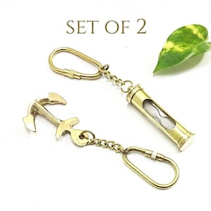 May include: A set of two gold-toned metal keychains featuring a nautical anchor charm and a miniature hourglass pendant. Each keychain includes a metal chain and a screw-lock carabiner attachment. The text SET OF 2 is printed above the items.