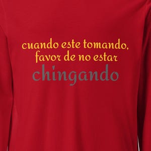 May include: A red long-sleeve t-shirt featuring the text "cuando este tomando, favor de no estar chingando" printed on the front. The first two lines of the text are in a yellow font, while the word "chingando" is in a grey font.