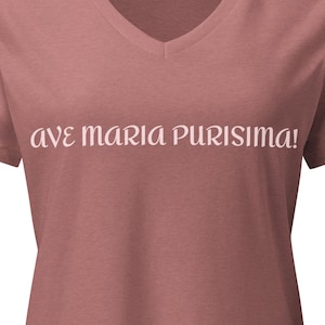 May include: A dusty rose V-neck t-shirt featuring the text "AVE MARIA PURISIMA!" printed in a light cream color across the chest. The garment has short sleeves and a standard fit, showcasing a solid color fabric with a clean, simple design.