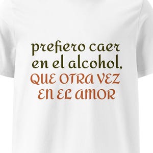 May include: A white short-sleeved t-shirt featuring dark green and reddish-brown text printed on the chest. The text reads: prefiero caer en el alcohol, QUE OTRA VEZ EN EL AMOR. The garment has a crew neckline and a standard straight hem.