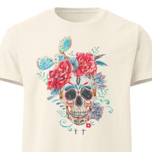 May include: A cream-colored short-sleeve t-shirt featuring a central graphic of a decorated skull adorned with vibrant red flowers, blue cactus, and green succulents. The skull design includes intricate colorful patterns and two small crosses below.