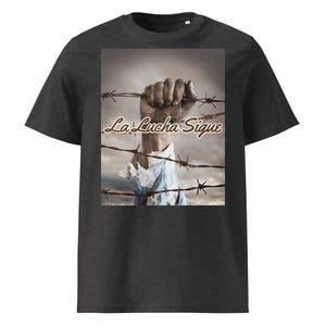 May include: A dark charcoal gray t-shirt featuring a rectangular graphic print of a clenched fist reaching upward through strands of barbed wire against a cloudy sky, with the text La Lucha Sique written in a brown script font across the center.