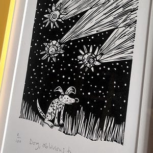 May include: Black and white linocut print of a dog looking up at three stylized comets against a starry sky. The print is framed in a yellow wooden frame. The text "Dog, oblivions to comets" is below the image.