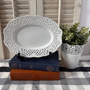May include: An oval white ceramic serving dish featuring a decorative lattice-work border with a scalloped edge. The dish is displayed on a black stand, resting atop a stack of two hardcover books, one blue and one brown, on a checkered surface.