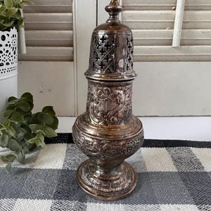 May include: A silver-toned metal sugar shaker featuring an ornate embossed floral and scroll pattern on the body. The removable lid has a perforated cross-shaped design and a small finial at the top, resting on a circular pedestal base.