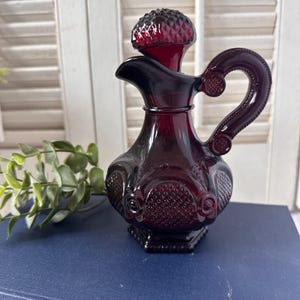 May include: A deep red glass cruet featuring a textured diamond pattern on the body and a matching stopper. The vessel has a curved handle, a wide spout, and a hexagonal base, showcasing a rich, translucent ruby colour throughout the glass structure.