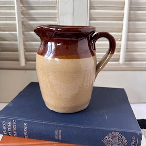 May include: A small ceramic pitcher featuring a two-tone design with a glossy dark brown glaze on the upper portion and a matte tan finish on the lower body. The pitcher has a curved handle and a small pouring spout, showcasing a simple, functional form.