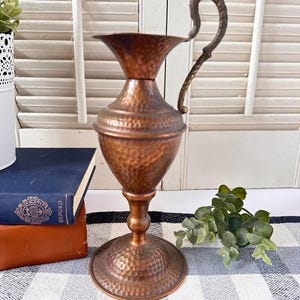 May include: A tall, hammered copper pitcher with a flared rim, a rounded body, and a pedestal base. The pitcher features a curved, textured metal handle attached to the side. The surface has a distinct dimpled, metallic finish throughout.