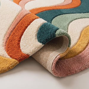 Op de afbeelding: Een getuft karpet met een golvend, veelkleurig patroon in tinten cr&egrave;me, gebrand oranje, mosterdgeel, saliegroen, diep petrol en oudroze. Het kleed heeft een dikke, getextureerde pool en een zichtbare geweven achterkant waar het is omgevouwen.