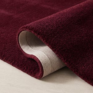 Può includere: Un tappeto color bordeaux intenso con una consistenza a pelo spesso e morbido. Un angolo del tappeto è ripiegato per rivelare il retro in tessuto intrecciato di colore chiaro, che presenta un motivo a griglia visibile sulla parte inferiore.