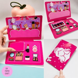 Puede incluir: Un kit de maquillaje de papel rosa que presenta varios cosméticos ilustrados como lápiz labial, pinceles y paletas. El conjunto incluye un estuche plegable con una silueta de princesa en la cubierta y múltiples compartimentos para organizar accesorios.