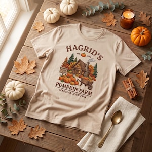 Puede incluir: Una camiseta beige de manga corta con un gráfico de una cabaña, calabazas y un carro de madera. El texto HAGRID'S est. 1692, PUMPKINS POTIONS & PLANTS, PUMPKIN FARM y MAGICALLY GROWN aparece en letras marrones en la parte delantera.
