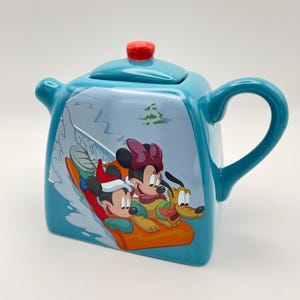 May include: A square-shaped turquoise ceramic teapot featuring a graphic of Mickey Mouse, Minnie Mouse, and Pluto riding an orange sled on snow. The teapot has a curved handle, a short spout, and a lid topped with a small, round, bright red knob.