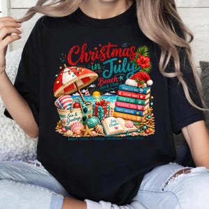 Puede incluir: Una camiseta negra con un estampado gráfico que incluye el texto 'Christmas in July Beach Book Club'. El diseño muestra una sombrilla de playa, un bolso, una pila de libros con títulos, un regalo y conchas marinas sobre arena.