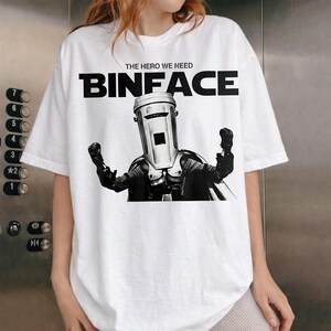 Puede incluir: Una camiseta blanca de manga corta con un gráfico en blanco y negro de una figura con un cubo de basura por cabeza, que lleva una capa y guantes. Sobre la figura, el texto dice THE HERO WE NEED y la palabra BINFACE en letras grandes y negritas.