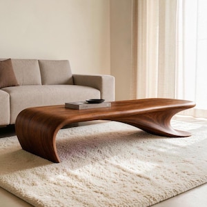 May include: A low-profile coffee table crafted from dark brown wood with a continuous, curved, wave-like silhouette. The surface is smooth with visible wood grain patterns, featuring a single rectangular book placed flat on the tabletop.