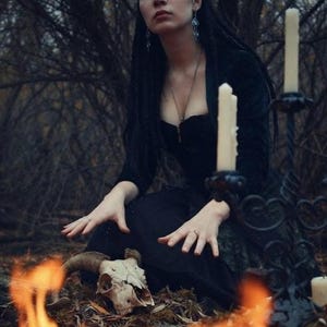 May include: A black dress with a sweetheart neckline, worn with a silver necklace and drop earrings. An animal skull with curved horns rests on the ground near a black metal candelabra holding three white pillar candles, with flames in the foreground.