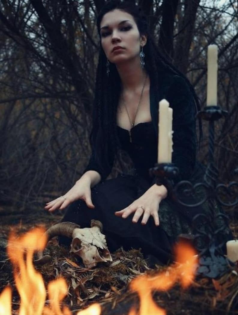 May include: A black dress with a sweetheart neckline, worn with a silver necklace and drop earrings. An animal skull with curved horns rests on the ground near a black metal candelabra holding three white pillar candles, with flames in the foreground.