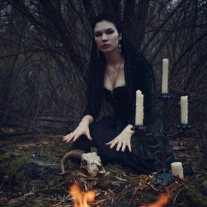 May include: A black candelabra with four white pillar candles, a ram skull with curved horns, and a person wearing a black long-sleeved dress with a corset-style bodice, positioned in a dark forest setting with a small fire in the foreground.