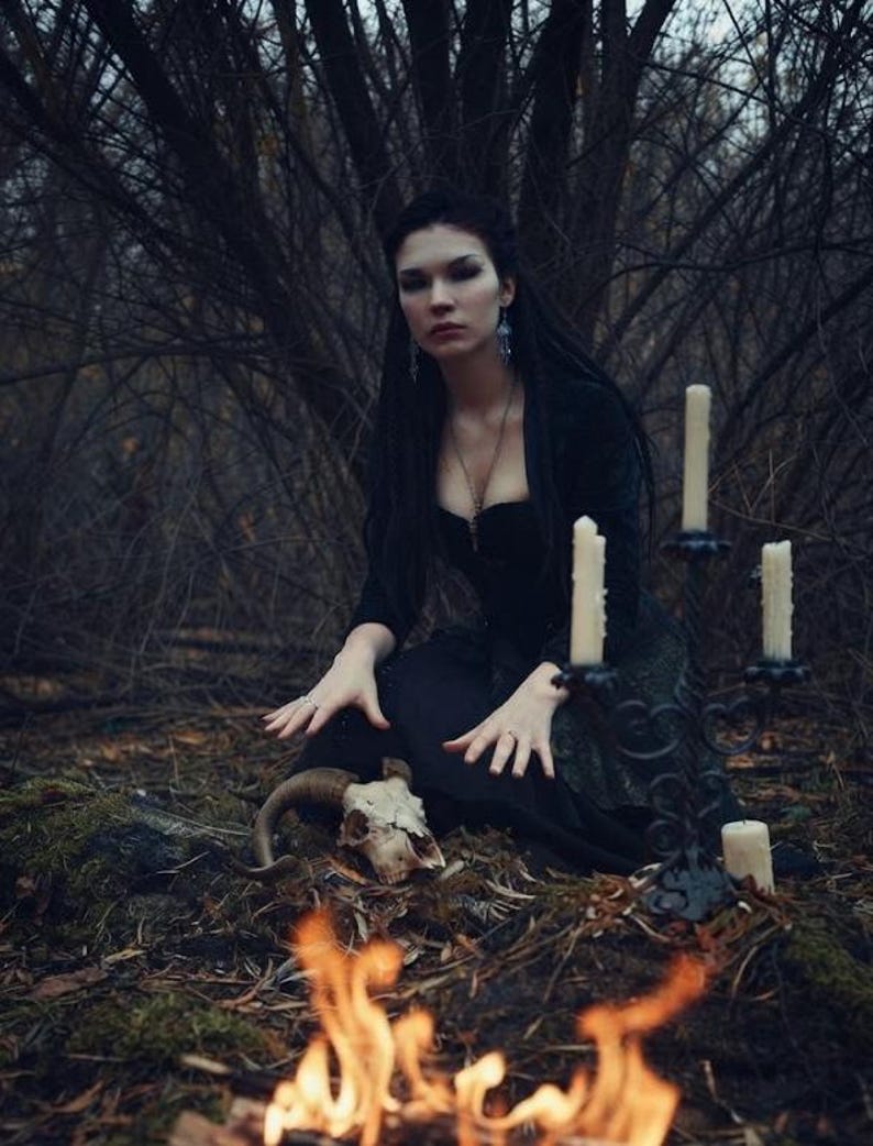 May include: A black candelabra with four white pillar candles, a ram skull with curved horns, and a person wearing a black long-sleeved dress with a corset-style bodice, positioned in a dark forest setting with a small fire in the foreground.