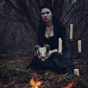 May include: A ram skull with curved horns held in front of a person wearing a black long-sleeved top and a dark skirt. Beside them stands a black metal candelabra holding three off-white pillar candles, with flames visible in the foreground.