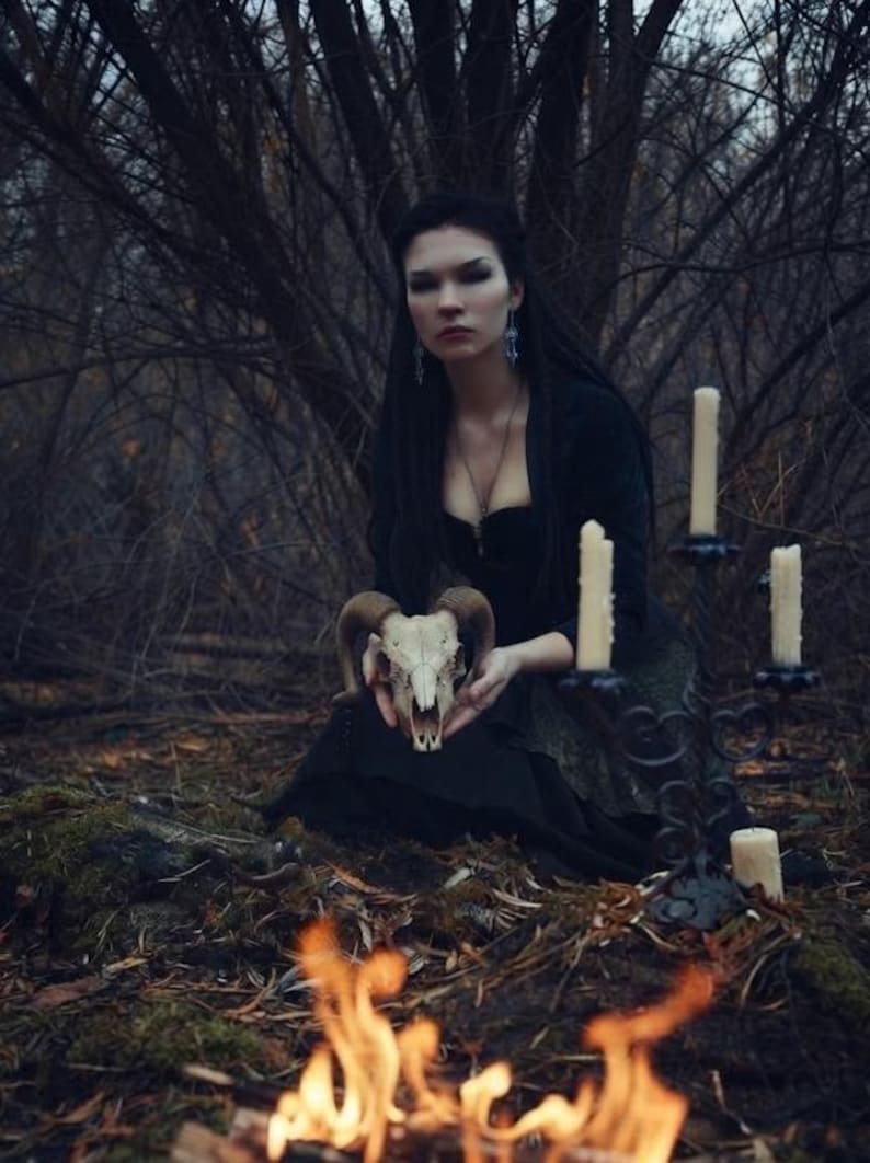 May include: A ram skull with curved horns held in front of a person wearing a black long-sleeved top and a dark skirt. Beside them stands a black metal candelabra holding three off-white pillar candles, with flames visible in the foreground.