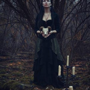 May include: A long black dress with a layered skirt and a fitted bodice, worn with a dark long-sleeved top. The attire is paired with a ram skull held in the hands and a black metal candelabra featuring four white pillar candles nearby.