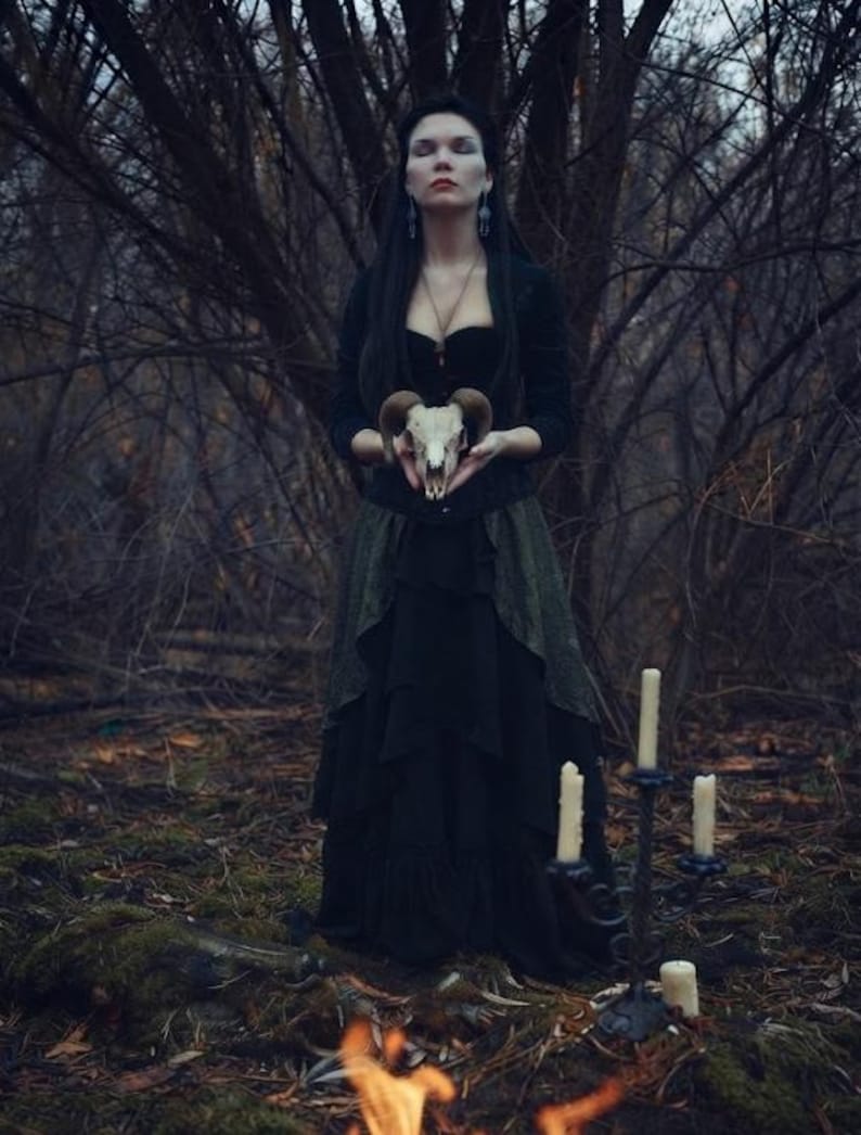 May include: A long black dress with a layered skirt and a fitted bodice, worn with a dark long-sleeved top. The attire is paired with a ram skull held in the hands and a black metal candelabra featuring four white pillar candles nearby.
