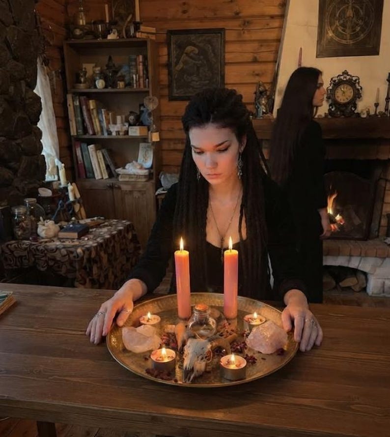 May include: A large round brass tray holding two tall pink pillar candles, four small tea light candles, two large raw quartz crystals, a small glass bottle, and an animal skull, all arranged on a wooden table in a rustic room.