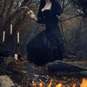 May include: A black corset dress with a layered skirt, worn with a dark bolero jacket. The attire features intricate dark detailing and a fitted bodice. The ensemble is paired with silver drop earrings and a delicate necklace, creating a cohesive look.