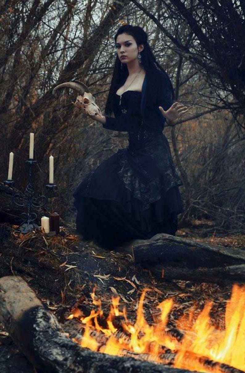 May include: A black corset dress with a layered skirt, worn with a dark bolero jacket. The attire features intricate dark detailing and a fitted bodice. The ensemble is paired with silver drop earrings and a delicate necklace, creating a cohesive look.