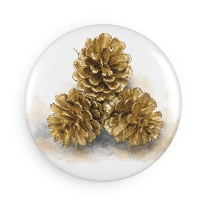 May include: A circular button featuring a watercolor illustration of three metallic gold pinecones arranged in a triangular cluster. The artwork is set against a soft, neutral-toned wash background, highlighting the textured scales of the pinecones.